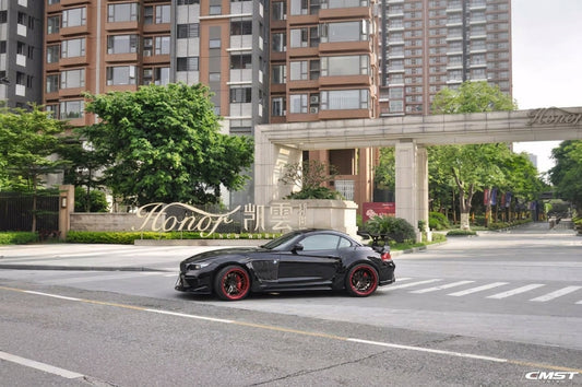 Black BMW Z4 roadster wearing the CMST widebody carbon fibre kit with red-barrel two-piece forged wheels, front three-quarter low angle with visible carbon weave