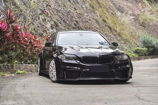 BMW 5 Series F10 in gloss black wearing the CMST carbon widebody aero kit, front three-quarter low angle with visible carbon-weave lip and flared arches
