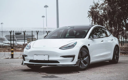 White Tesla Model 3 wearing a CMST carbon fibre body kit with front lip, side skirts and boot spoiler against a moody studio background
