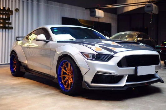 Silver 2015-2017 S550 Ford Mustang with the CMST widebody kit on 20-inch CMST CT201 2-piece forged wheels with a gold face and blue lip, red Brembo brakes behind, low front three-quarter studio angle