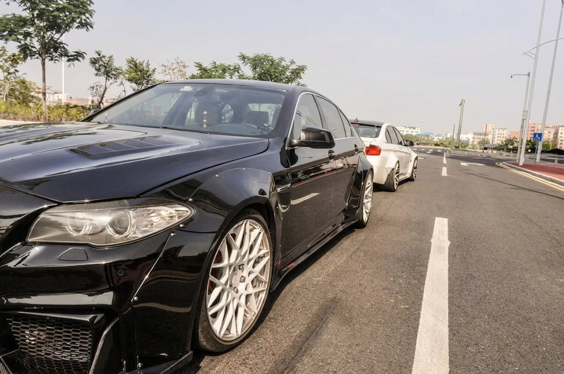 BMW 5 Series F10 in gloss black wearing the CMST carbon widebody kit, front three-quarter low angle with visible carbon-weave aero